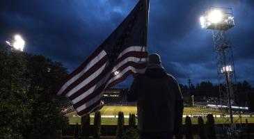 Photo taken behind a man holding an American flag looking down at a lit sportsfield at night